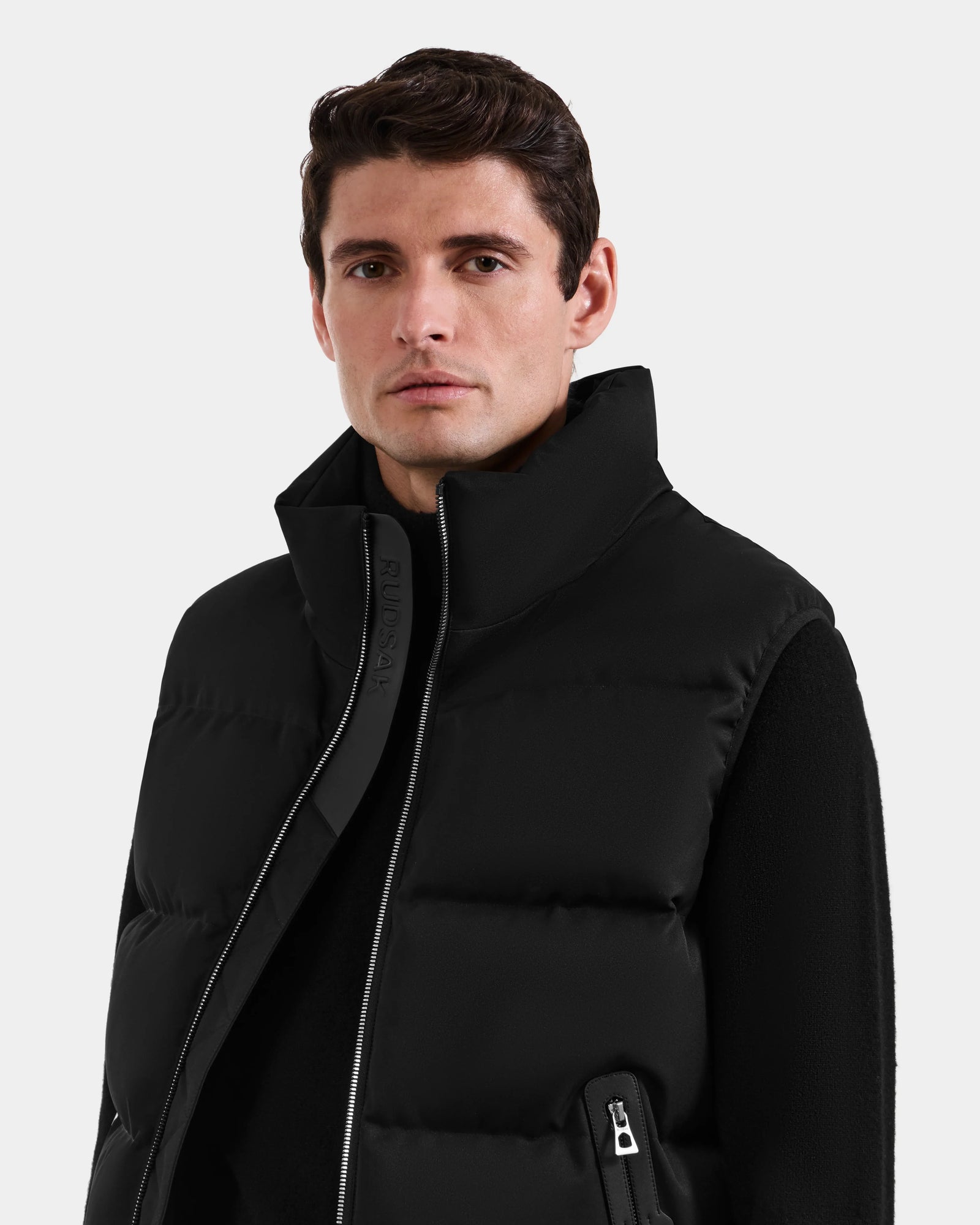 A man with short brown hair wears the Rudsak HARLOW - M125528 Black men’s puffer vest over a black long-sleeve shirt, standing against a plain light gray background and looking slightly to the side.