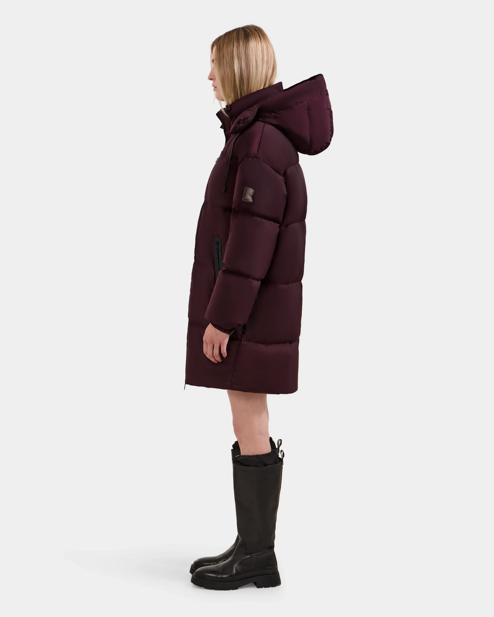A person in the Rudsak KARLEE L - 8125682 Bordeaux, a dark purple long down puffer coat with hood, stands in profile facing left. Paired with black knee-high boots, they showcase chic cold-weather style against a white background.
