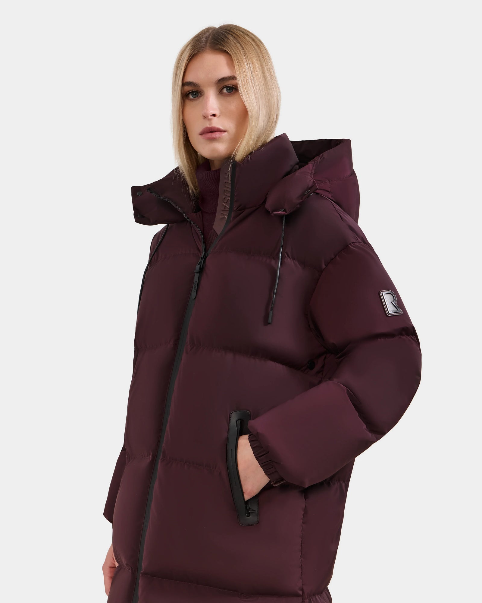 A woman wears the Rudsak KARLEE L - 8125682 Bordeaux maroon long down puffer jacket with a hood, black zipper, and sleeve patch, standing against a plain light background.