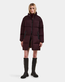A woman stands against a white background wearing the Rudsak KARLEE L - 8125682 Bordeaux long-length down puffer jacket, paired with brown knee-high boots and a neutral expression—ideal for cold-weather wear.