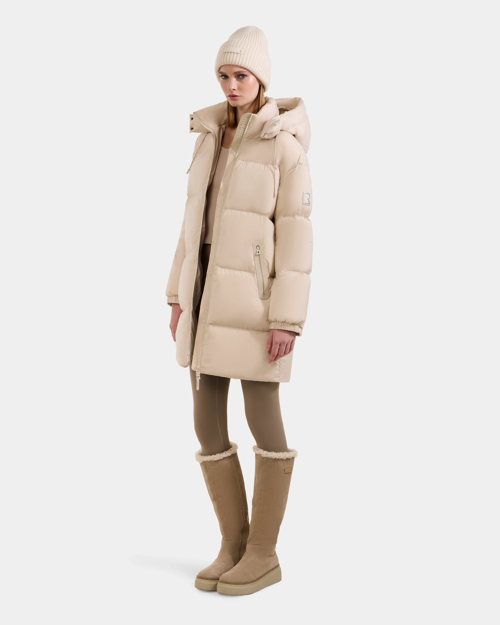 A woman stands against a white background wearing the Rudsak KARLEE L - 8125682 Vanilla, a beige long-length down puffer jacket with vegan leather trims, styled with a beige beanie, tan leggings, and tall tan boots.