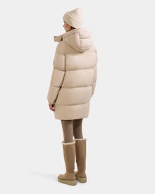 A person wearing the Rudsak KARLEE L - 8125682 Vanilla women’s long down puffer with vegan leather trims, tan leggings, beige boots, and a beige knit hat stands facing away against a white background. This jacket is stylish and cozy.