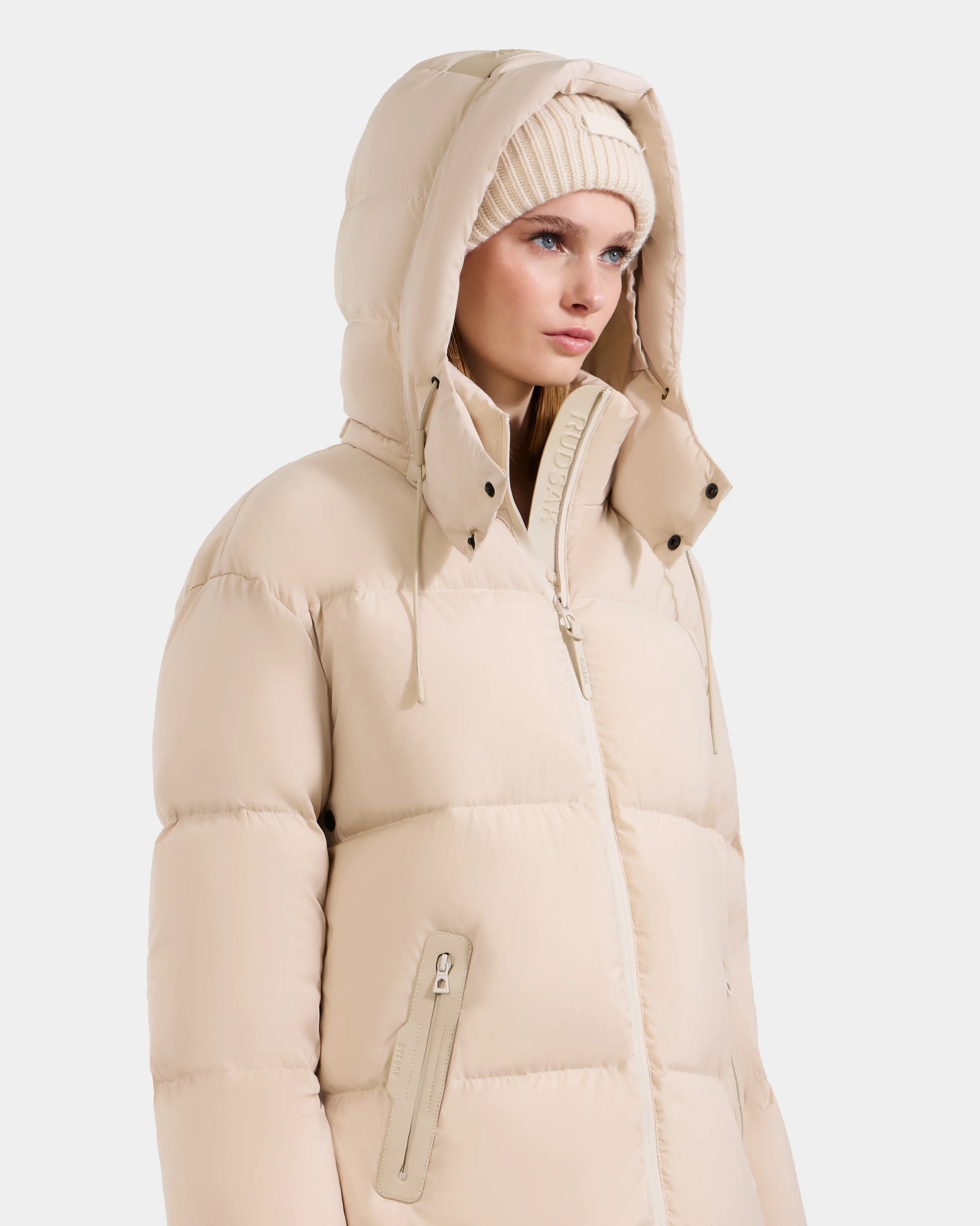 A woman wearing the Rudsak KARLEE L - 8125682 Vanilla long down puffer with vegan leather trims and a matching knit beanie, looking slightly to the side against a plain white background.