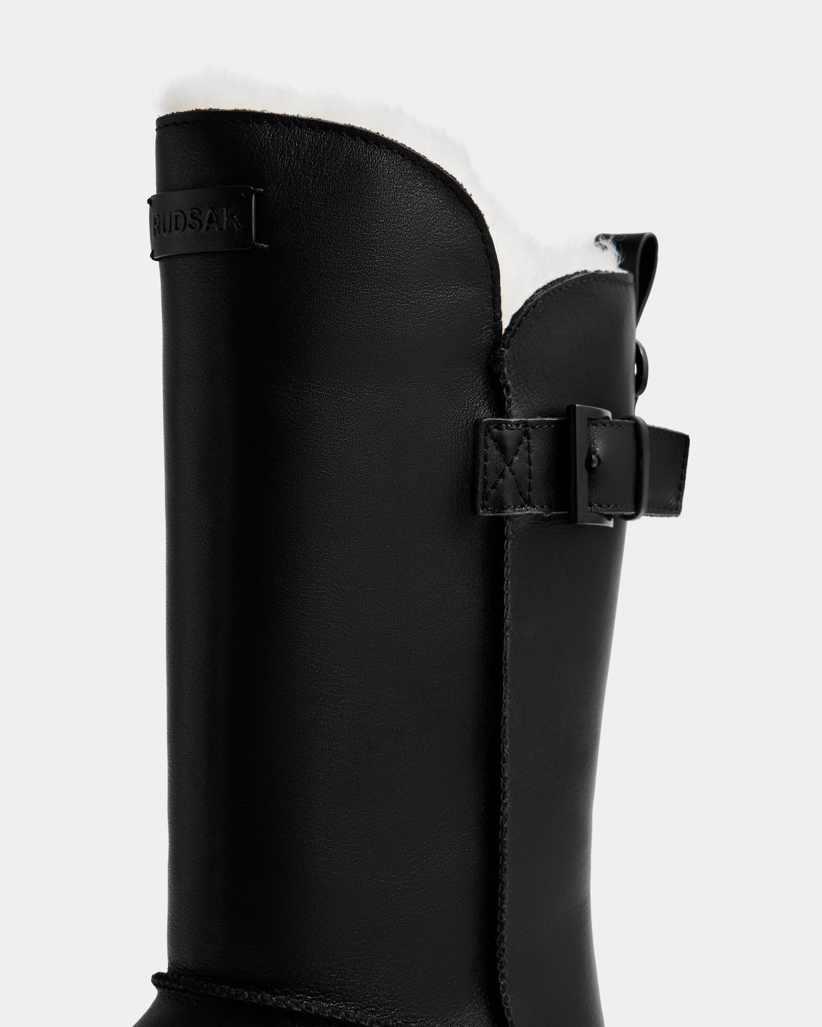 Close-up of the Rudsak KLAIR L - L225519 Black women’s leather boot featuring a white wool-lined interior, side strap with buckle, and brand label near the top—ideal city boots set against a plain gray background.
