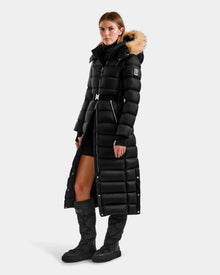 A woman wears the Rudsak LILITH L - L125528 Black puffer coat with a fur-trimmed hood over a black turtleneck dress and knee-high boots, standing against a plain light background.