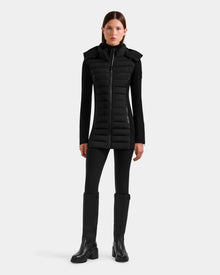 A woman faces forward in the Rudsak MARRY M - L125519 Black women's mid-length hybrid coat, layered over a black top, pants, and knee-high boots against a plain white background.