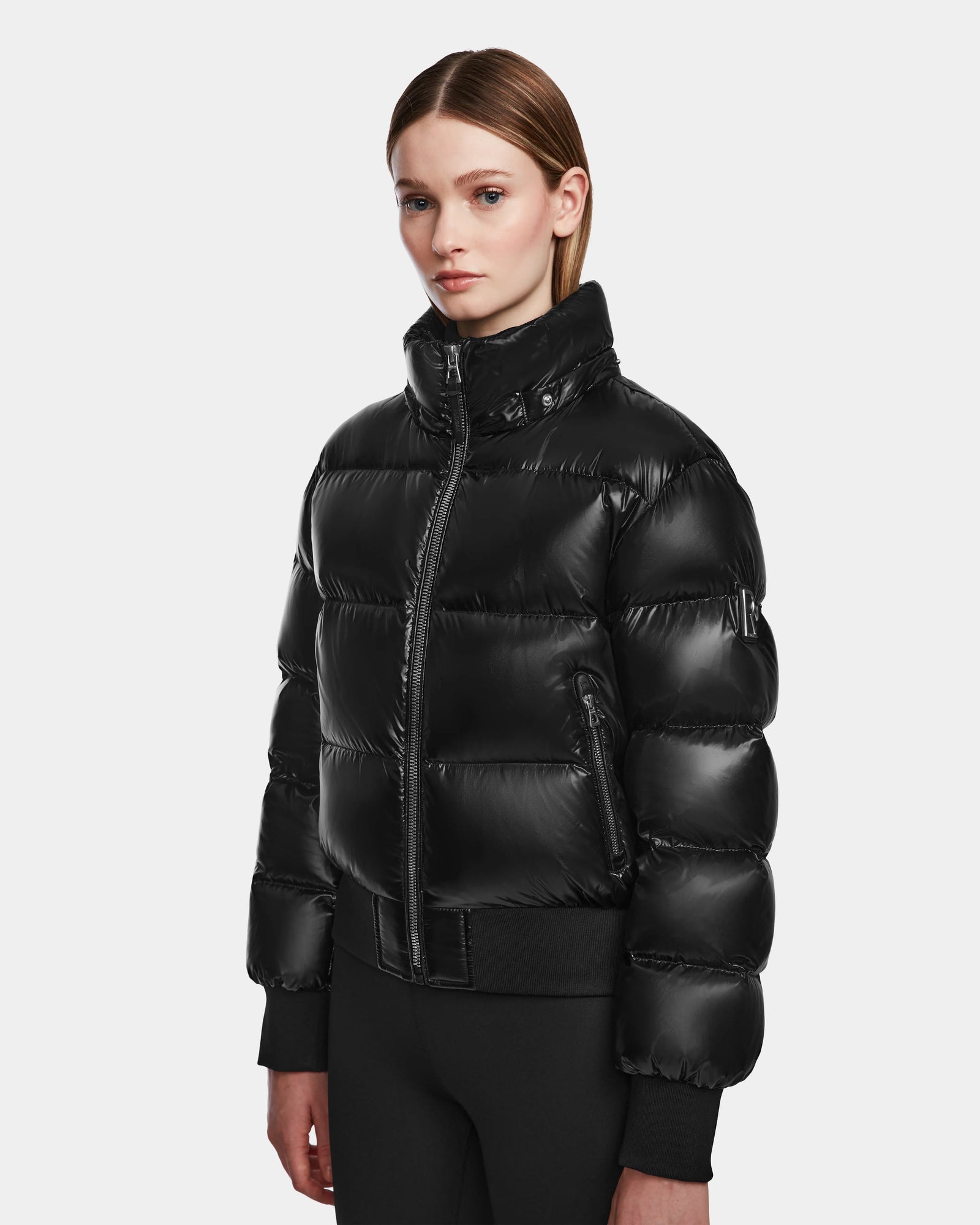 A woman with straight, shoulder-length hair wears the Rudsak MAUDE Q-S - L124553 Black down jacket, featuring a shiny finish, high collar, and zippered front, while standing against a plain white background.