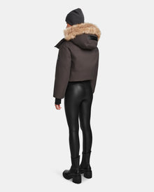 Person standing with their back to the camera, wearing the Rudsak MAUDE - L124522 Grey winter jacket with a fur-lined hood, paired with black faux-leather leggings, black boots, and a dark beanie, set against a plain white background.