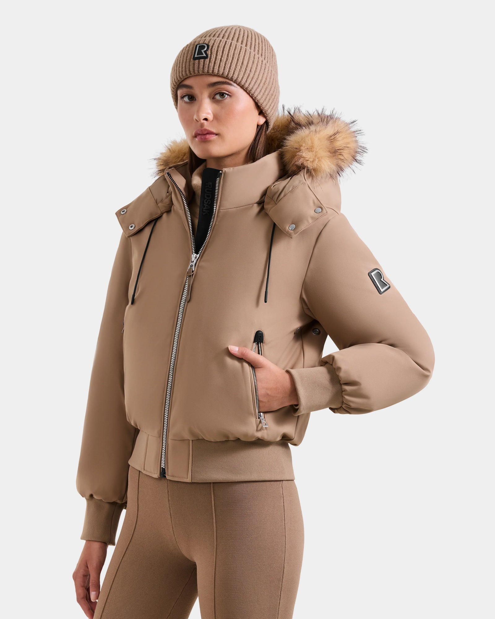 A woman wears the Rudsak MAUDE - L124522 Sand winter jacket with premium down and a fur-trimmed hood, paired with a matching ribbed knit beanie and fitted ribbed leggings, standing against a plain light background.