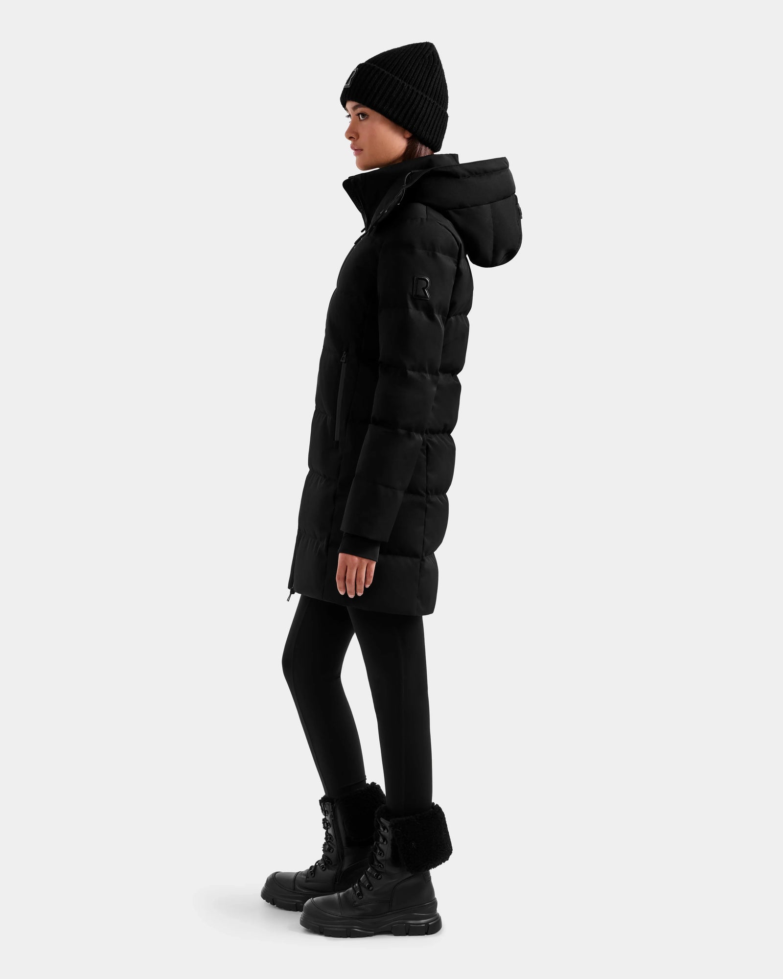 A woman in the Rudsak MIRIAM MW - L125604 Black down puffer, black leggings, boots, and a beanie stands sideways against a plain light gray background.