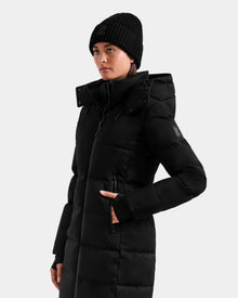 Wearing the Rudsak MIRIAM MW - L125604 Black women’s down puffer coat with a hood and a black knit beanie, she stands against a plain light background, looking slightly to the side with one hand in her pocket.
