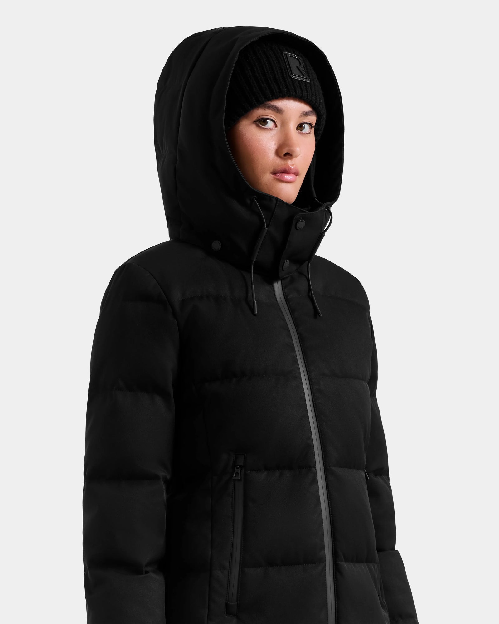 A person wearing the Rudsak MIRIAM MW - L125604 Black down winter coat and a black knit beanie stands against a plain light gray background.