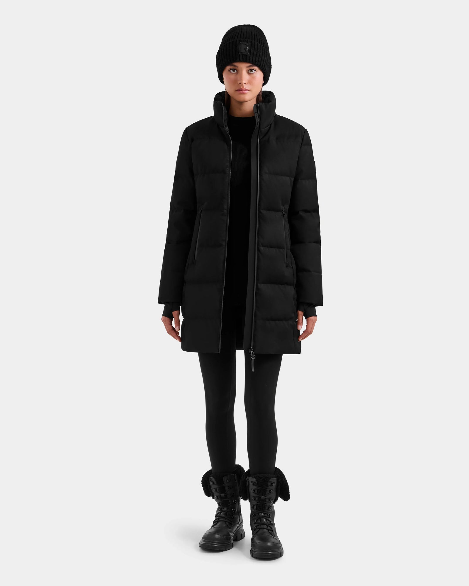 A person faces forward wearing the Rudsak MIRIAM MW - L125604 Black women’s down puffer coat with RDS-certified down, styled with a black beanie, leggings, and boots against a plain light gray background.
