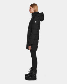 A woman stands in profile wearing the Rudsak MIRIAM - L123544 Black womens down puffer jacket with a visible sleeve logo, paired with black leggings and winter boots against a plain white background.