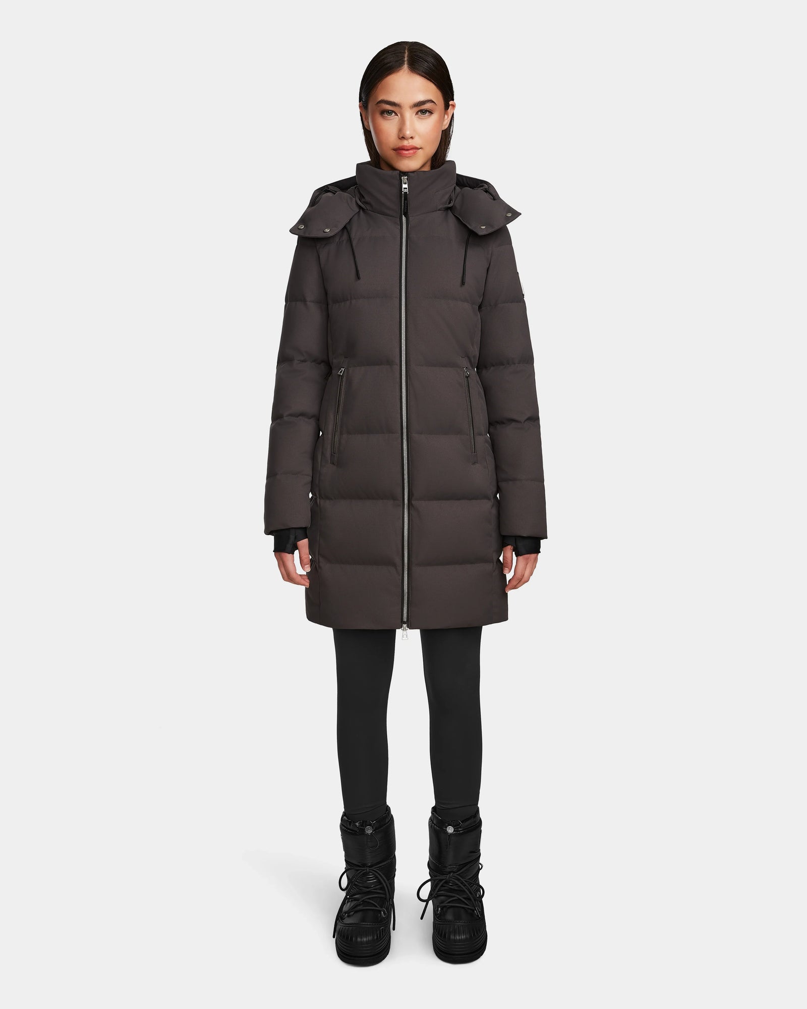 A woman stands facing forward, wearing the Rudsak MIRIAM - L123544 Grey womens down puffer coat with a hood, paired with black leggings and winter boots against a plain white background—a stylish pick for winter.