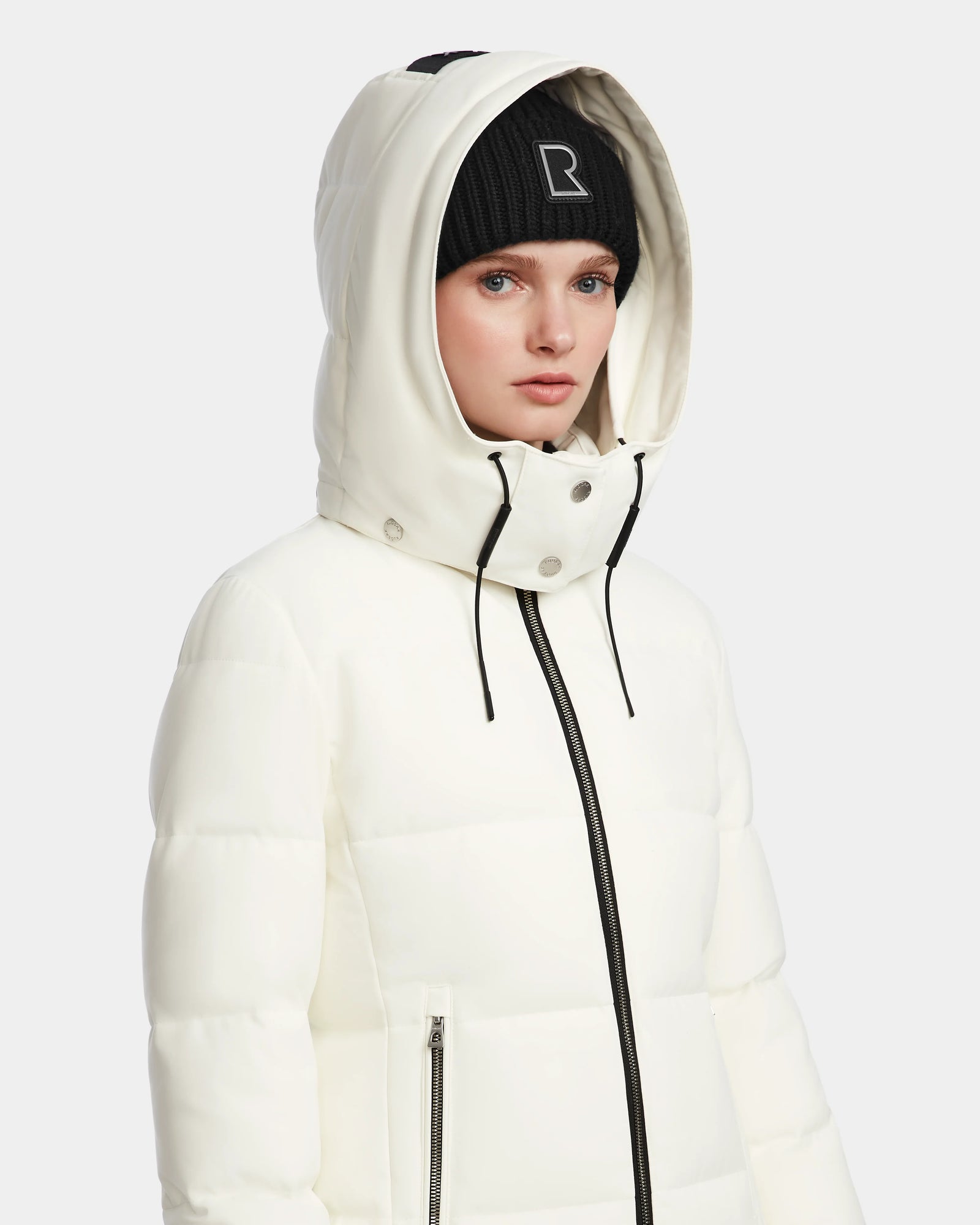 A woman wears the Rudsak MIRIAM - L123544 Iceland white RDS-certified down puffer jacket with the hood up and a black “R” logo beanie, facing forward with a neutral expression against a plain light background.