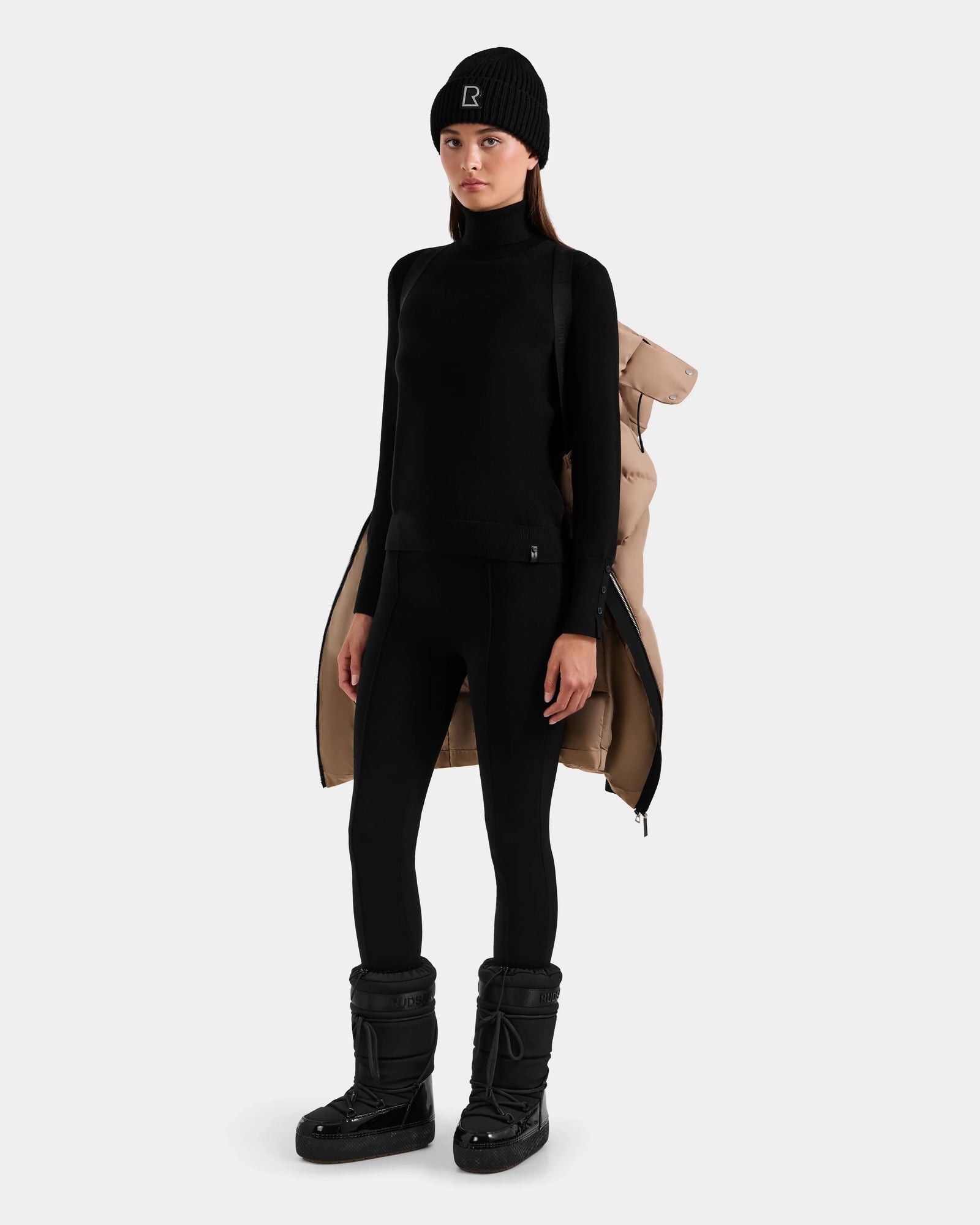 A woman stands facing forward, wearing a black beanie, turtleneck, leggings, snow boots, and the beige Rudsak MIRIAM - L123544 Sand women’s down puffer jacket draped over her shoulders against a plain light background.
