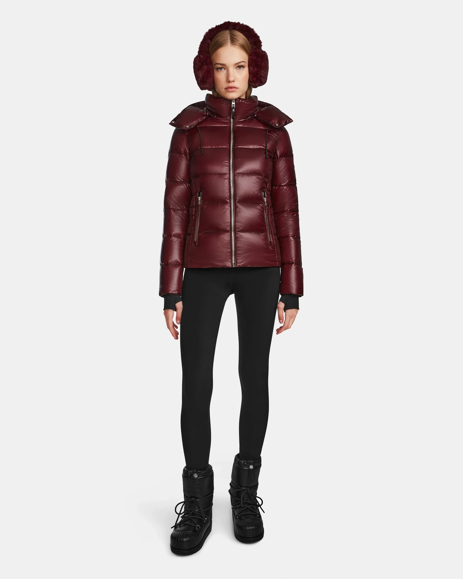 A woman faces forward in a shiny wine Rudsak MIRRY S - L123545 womens quilted down coat, with matching earmuffs, black leggings, and black winter boots, standing against a plain white background.