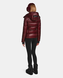 A woman in the Rudsak MIRRY S - L123545 Wine puffer jacket, black leggings, black winter boots, and maroon earmuffs stands with her back to the camera against a plain white background.