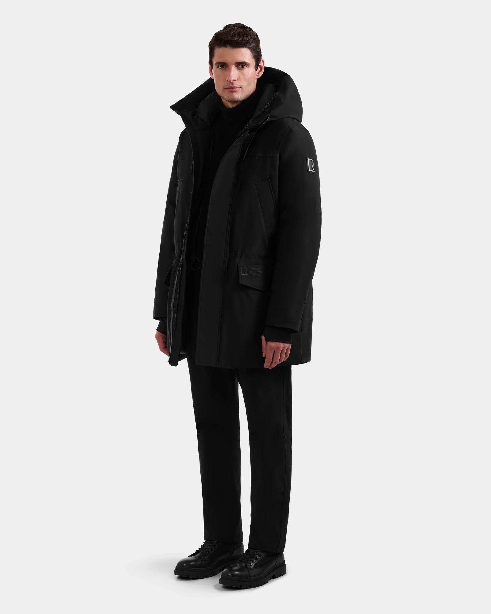 A man faces forward against a light background, wearing the Rudsak MITCHELL - M125513 Black RDS-certified down winter parka, paired with black pants, shoes, and top. His arms rest relaxed at his sides.