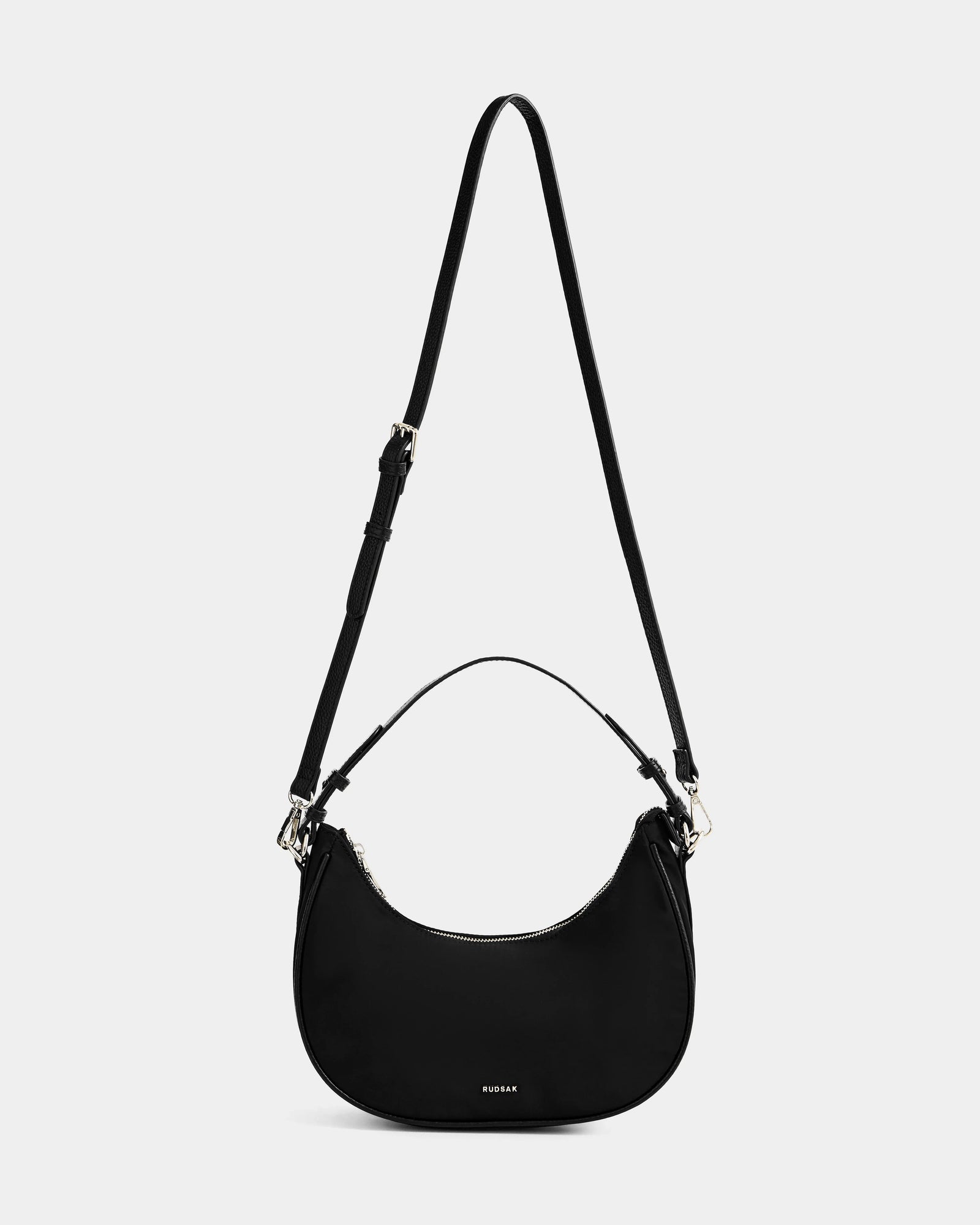 The Rudsak NIKKI - L325512 Black is a women's crescent shoulder bag with a hobo silhouette, short handle, adjustable crossbody strap, silver hardware, and small logo near the bottom, shown on a plain white background.