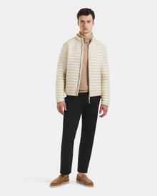 A man stands against a plain background wearing the Rudsak REYMOND M - M125523 Vanilla men’s down jacket, made with RDS-certified down, paired with a beige turtleneck sweater, black pants, and brown loafers.