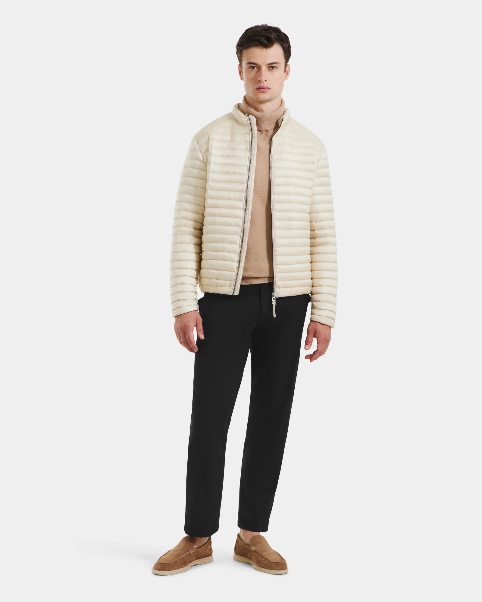 A man stands against a plain background wearing the Rudsak REYMOND M - M125523 Vanilla men’s down jacket, made with RDS-certified down, paired with a beige turtleneck sweater, black pants, and brown loafers.