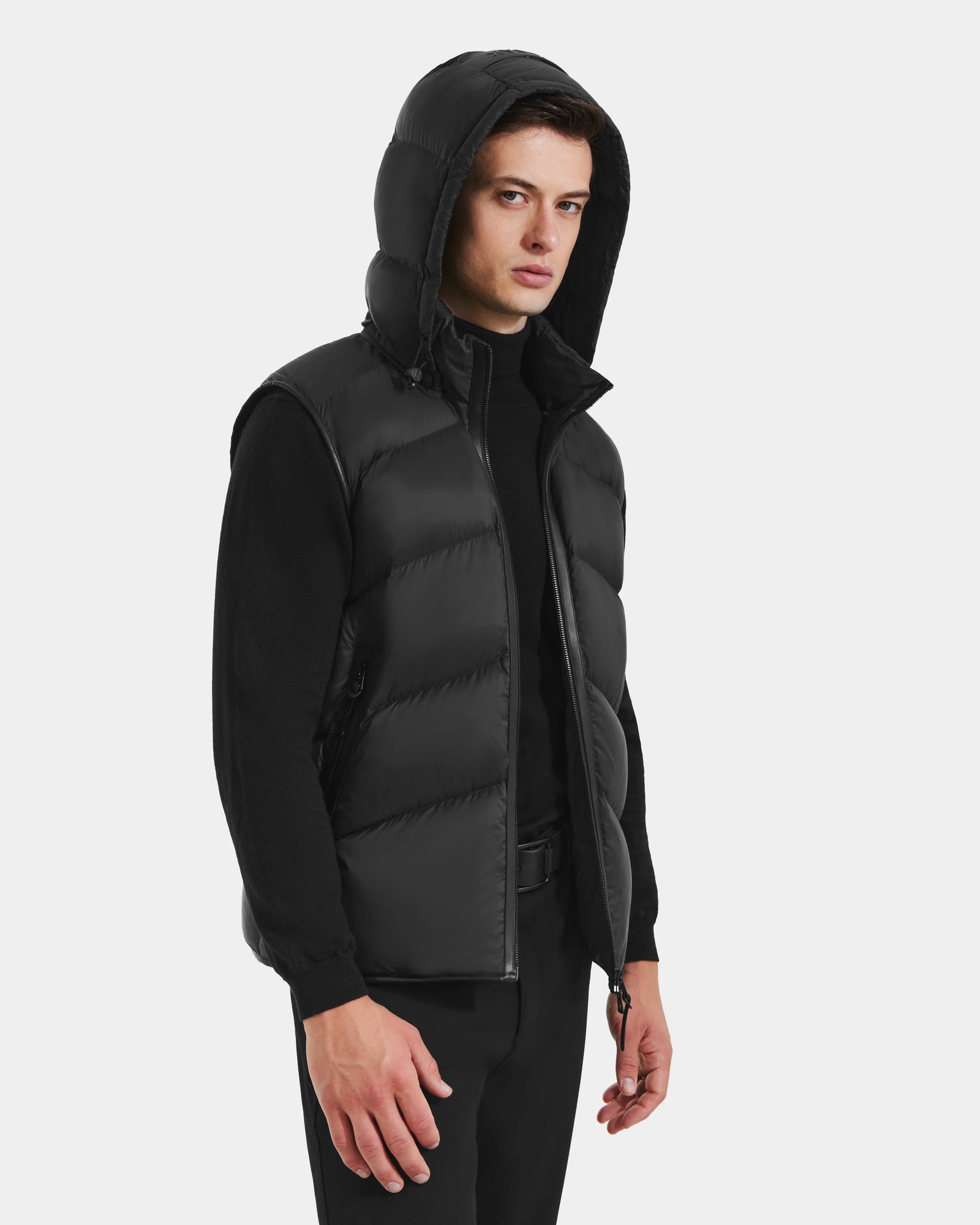 A man models the Rudsak RICHARDSON - M125527 Black men’s hooded vest, layered over a black sweater and pants, standing against a light background and highlighting its lightweight, water-repellent style.