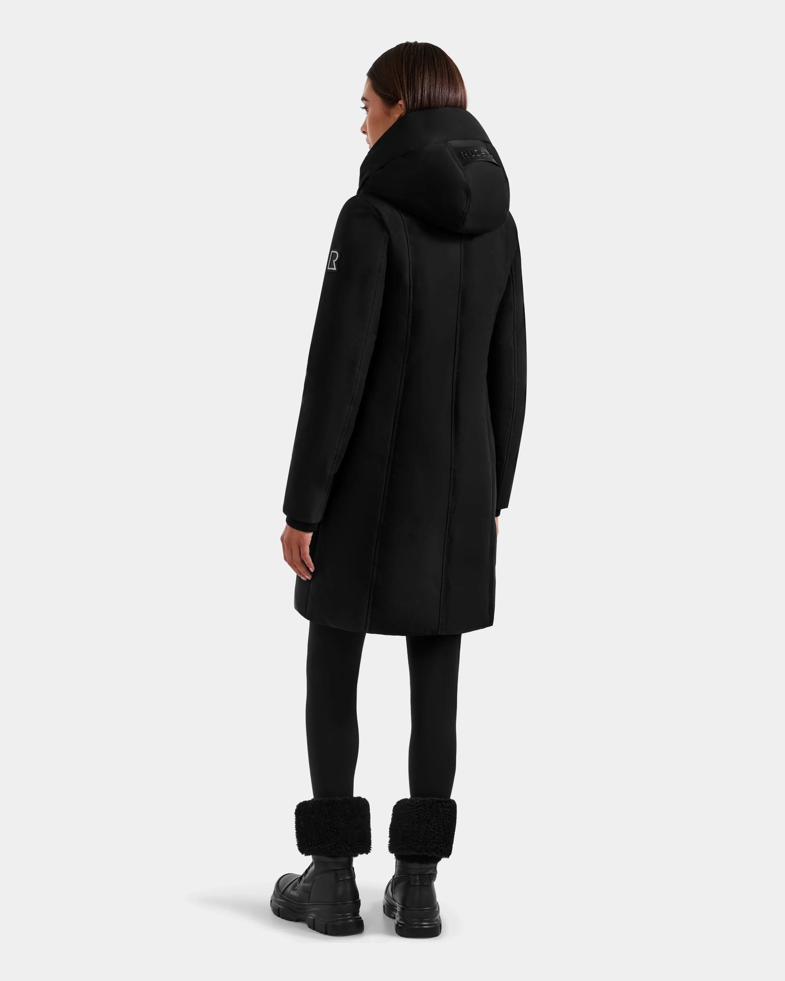A woman with straight brown hair faces away, wearing the SARINA - L125550 Black parka by Rudsak, paired with black leggings and black fur-cuff boots. The background is plain light gray.