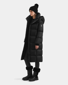 A person wears the Rudsak SASHA - 8125686 Black women’s long down puffer coat with a removable hood, paired with black leggings, fur-trimmed black boots, and a pom-pom beanie against a plain white background.
