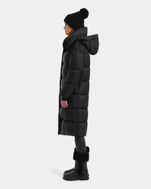 A person in the Rudsak SASHA - 8125686 Black women’s long down puffer coat with removable hood, black beanie with pom-pom, leggings, and boots stands in profile against a plain light background.