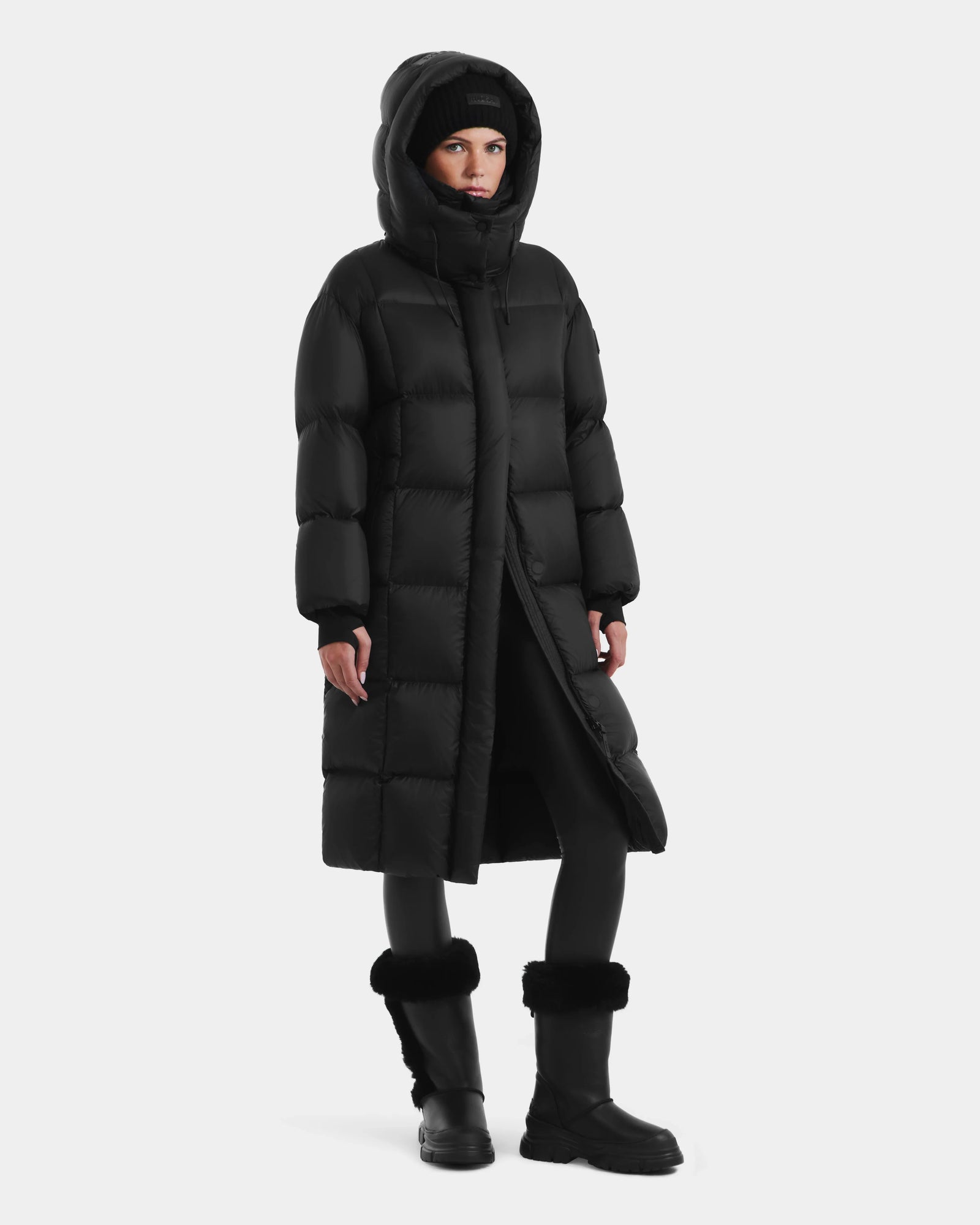 A woman stands against a light background wearing the Rudsak SASHA - 8125686 Black women’s long down puffer coat with removable hood, paired with black leggings and fur-trimmed boots.