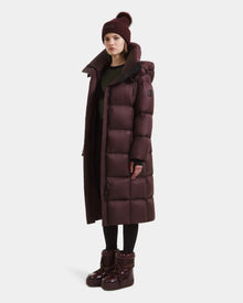 A woman wears the Rudsak SASHA - 8125686 Bordeaux women’s long down puffer coat with a high collar, paired with a matching beanie, black leggings, and shiny maroon boots, set against a plain white background.