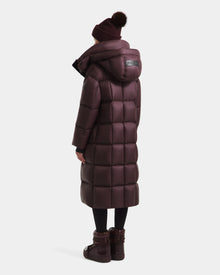 A person is seen from behind wearing the Rudsak SASHA - 8125686 Bordeaux women’s long down puffer coat with a hood, black leggings, a maroon pom-pom beanie, and matching boots.