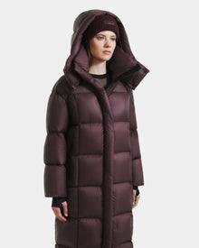A woman in the Rudsak SASHA - 8125686 Bordeaux long down puffer coat with a hood and a matching knit beanie stands facing forward against a plain white background.