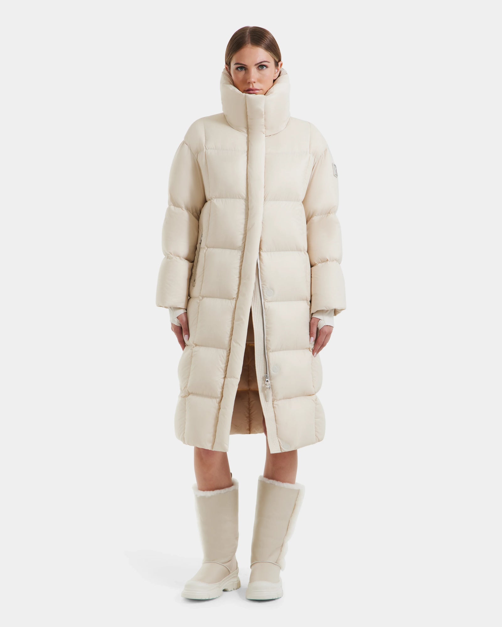 A woman faces forward wearing the Rudsak SASHA - 8125686 Vanilla long down puffer coat with a high collar and removable hood, paired with winter boots. A plain white background emphasizes the sleek cream style.