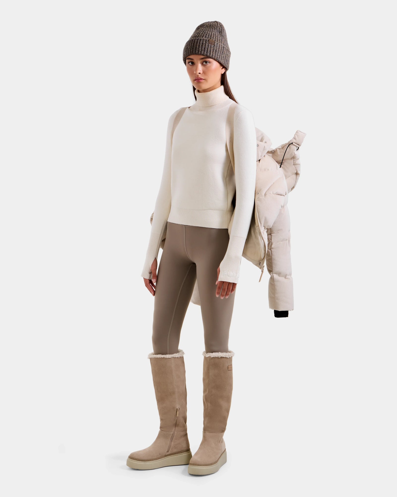 A woman stands against a white background wearing Rudsak’s STEFANIE V - L125584 Vanilla puffer over her shoulder, paired with a cream turtleneck, taupe leggings, beige knee-high boots, and a taupe beanie.