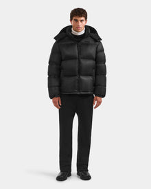 A person stands facing forward in a Rudsak STEFANO - 6125574 Black RDS-certified down jacket with a hood, paired with black pants, black shoes, and a white turtleneck top against a plain light gray background.