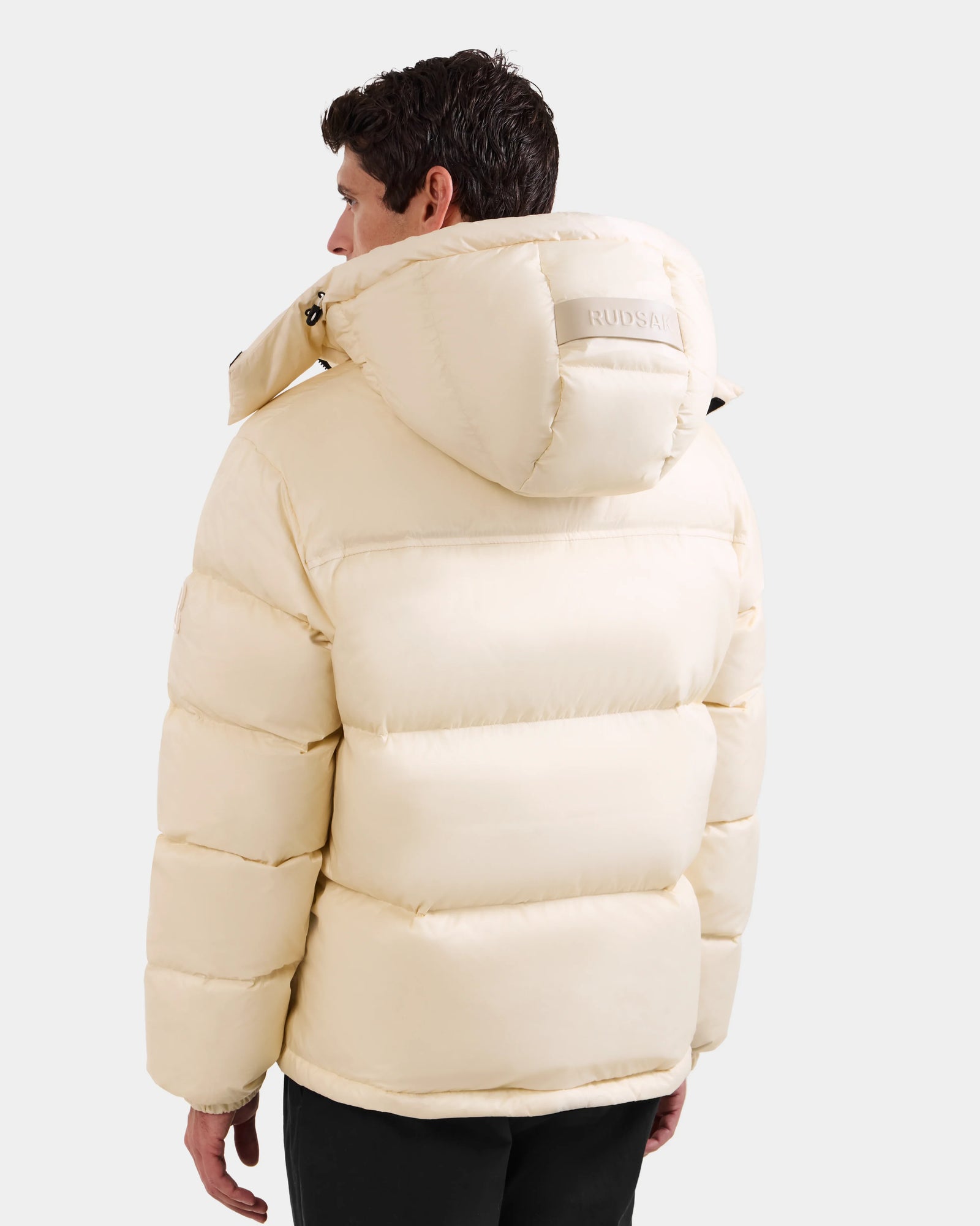 A man is seen from behind wearing the Rudsak STEFANO - 6125574 Vanilla men’s down puffer jacket with a hood. The light beige winter outerwear features horizontal stitching and a subtle brand logo on the hood, set against a plain background.