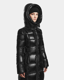 A person in the Rudsak VENICE S - L124514 Black shiny quilted down puffer coat with a hood and a black knit beanie stands against a plain light background, looking slightly to the side.