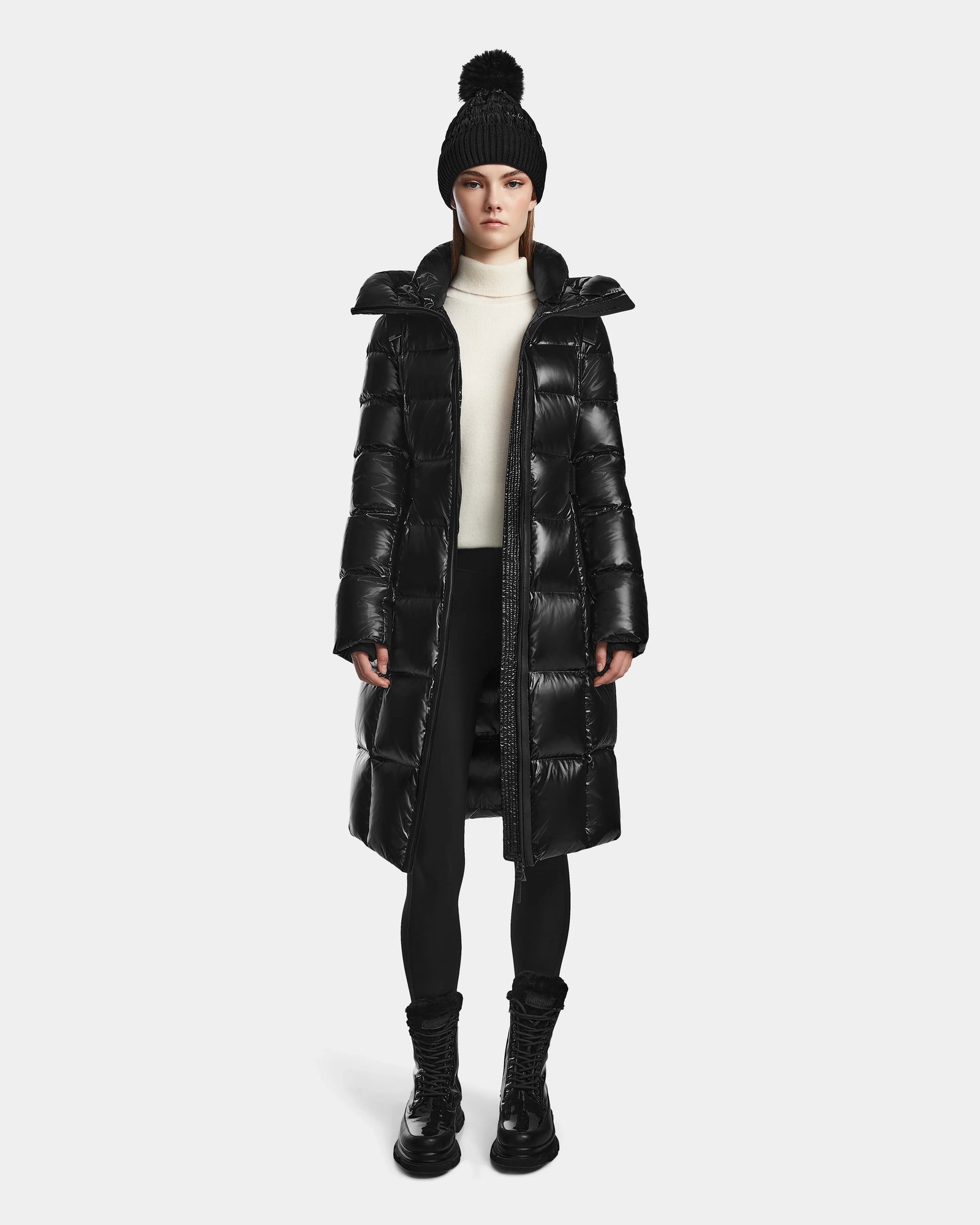 A woman wearing the Rudsak VENICE S - L124514 Black winter coat, a black pom-pom knit hat, white turtleneck, black leggings, and boots stands facing forward against a plain white background. The quilted puffer features RDS-certified down.