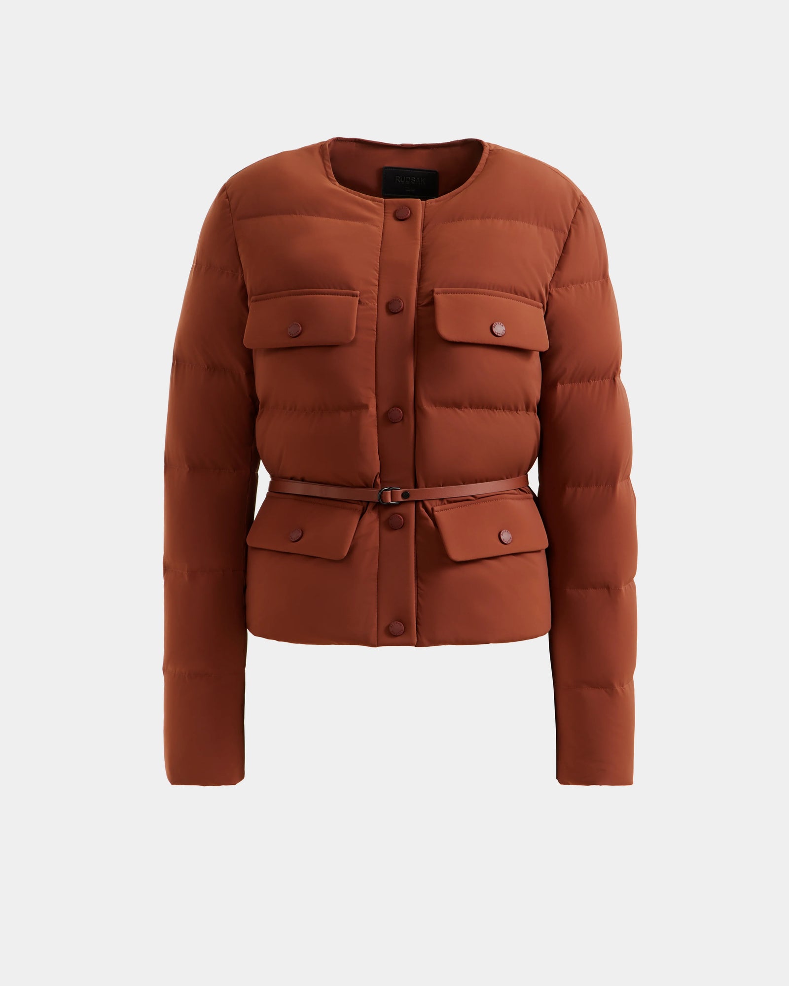 The Rudsak FRANCESCA - 8125021 Rust is a rust-colored quilted puffer jacket with a round neckline, button closure, chest and lower flap pockets, plus a matching vegan leather belt, shown on a plain white background.