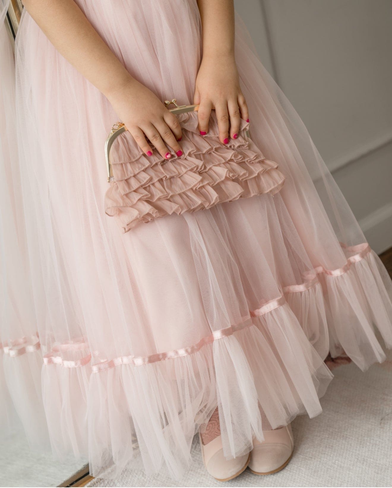 Girl holding dusty pink organza ruffle handbag against matching dress, featuring tiered ruffle detailing and gold metal frame