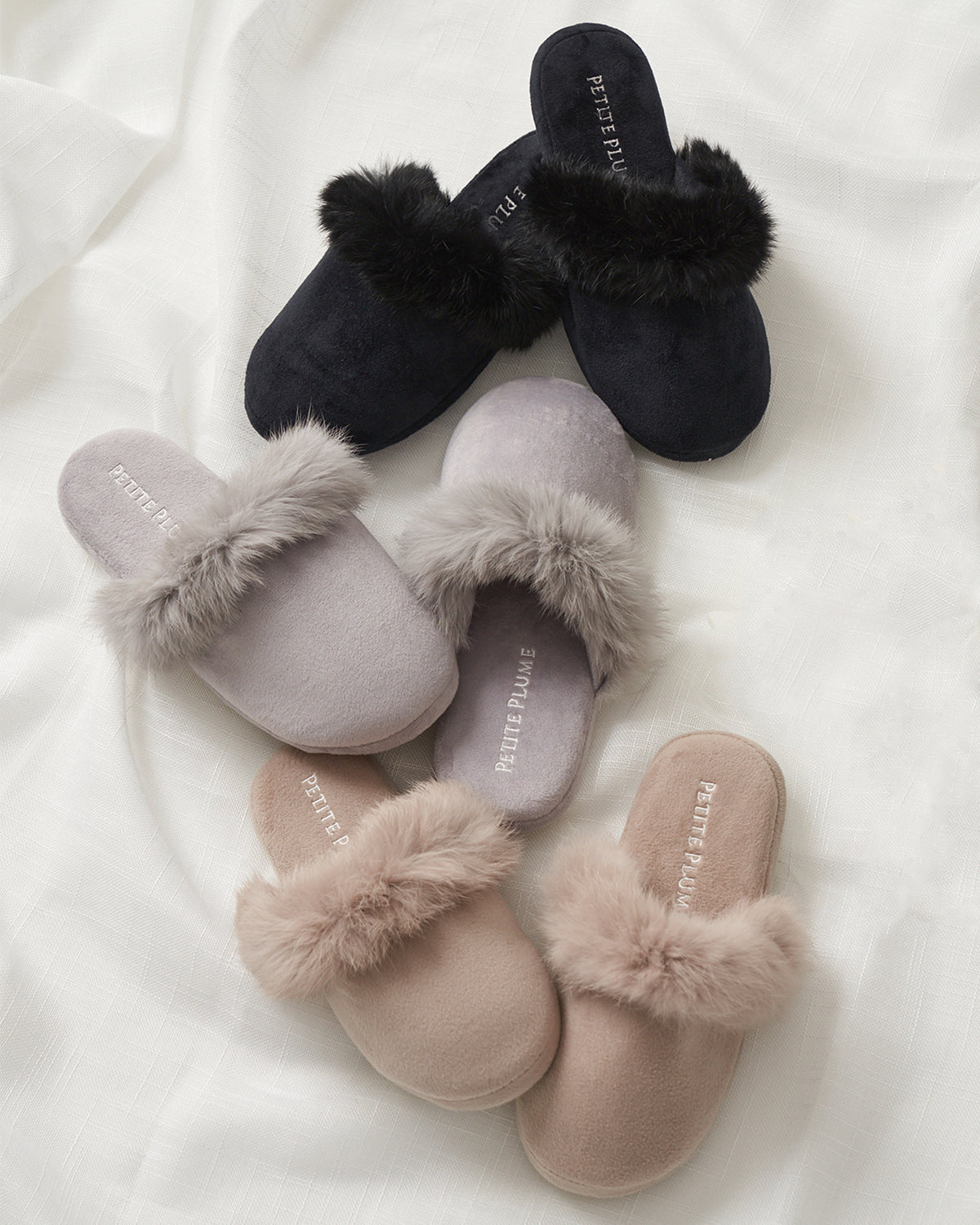 Three pairs of Petite Plumes Womens Faux Fur Trim Slippers in black, grey, and beige rest on a white fabric background. Ideal for any sleepwear collection, each features the brand name printed inside.
