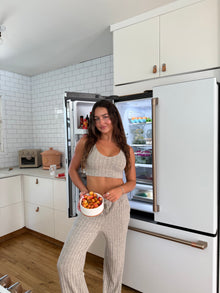 Woman wearing a beige ribbed knit loungewear set, featuring a cropped tank top with a scoop neckline and wide-leg drawstring pants with side pockets, standing barefoot against a plain white background.