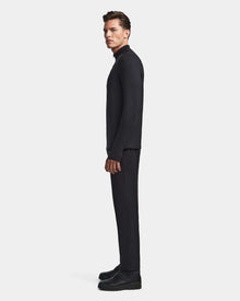 A man stands in profile against a plain light background, wearing the Rudsak WARREN - 6523502 Black zip-front mock neck mens fitted stretch top, along with matching black pants and black shoes.