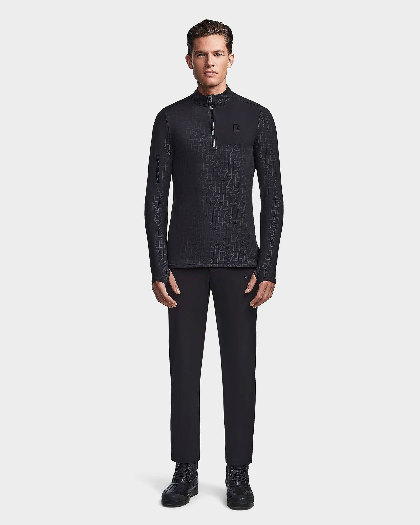 A man faces forward, wearing the Rudsak WARREN - 6523502 Black Pattern fitted stretch top—a black zip-front mock neck long-sleeve shirt—paired with black trousers and black lace-up shoes against a plain white background.