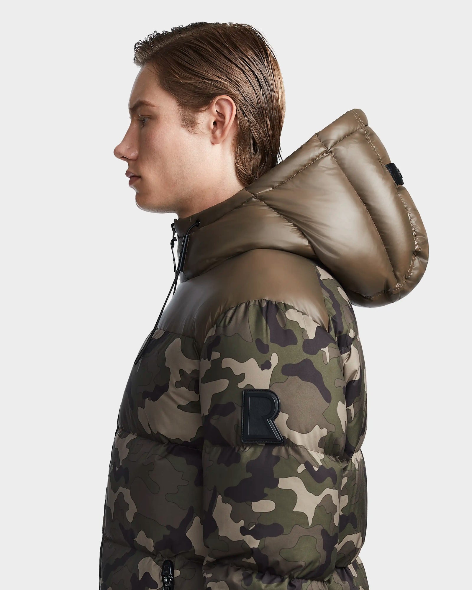 A person with light brown hair wears the Rudsak RONNY M - M123512 Camouflage mens puffer jacket, featuring a hood and black R patch on the upper sleeve. The weather-resistant outerwear is shown from the side on a plain white background.