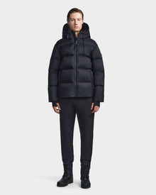 A person with short hair stands against a plain background, facing forward with arms relaxed, wearing the Rudsak DANIEL M - M123510 BLACK mens coat with a hood, paired with black pants and black boots.