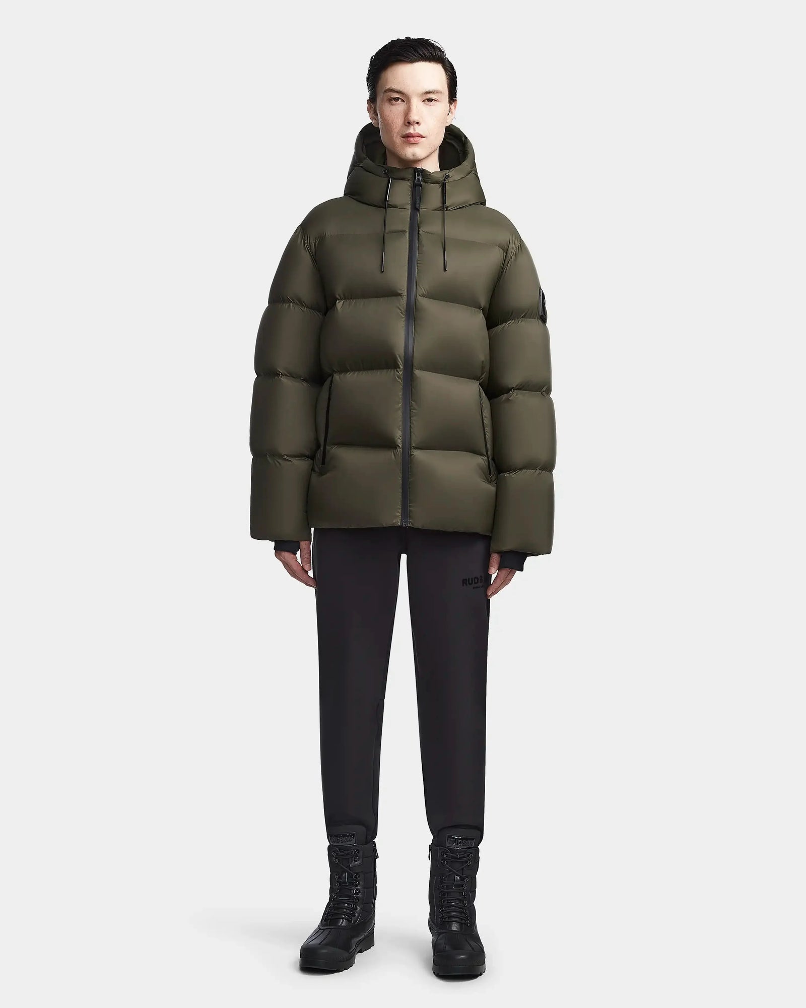 A person faces forward in a Rudsak DANIEL M - M123510 Light Olive weather-resistant winter jacket with hood, paired with black pants and black lace-up boots, set against a plain light background.